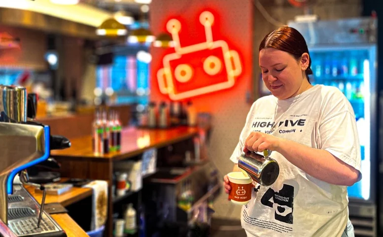 A Bar Robo barista pouring milk into a coffee beneath the neon robot sign.